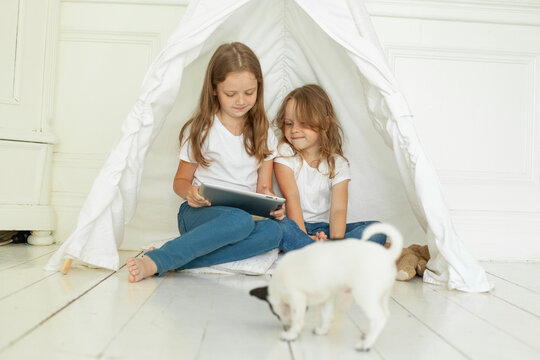 Cheerful little girl sisters with tablet pc computer in kids tent at home. Childhood, technology and hygge concept