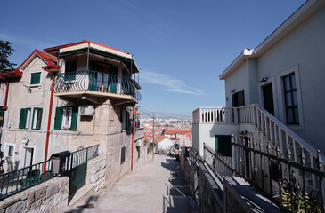 A narrow street in a Mediterranean town with traditional houses and a view of the sea in the background. The street is lined with stone buildings and balconies, under a clear blue sky.
