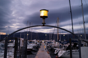 A marina at dusk with a focus on a lit lamp post in the foreground. The background features...