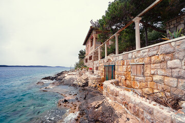 A rustic stone house by the sea with a rocky shoreline and clear blue water, surrounded by lush...