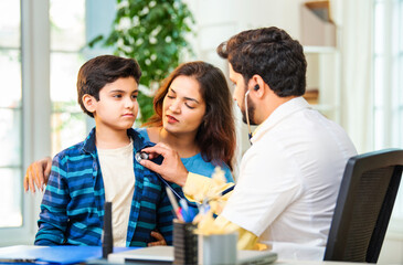 Male doctor checking boy’s symptoms with stethoscope as mother watches in hospital clinic setting