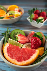 Close-up of bowl with fruits, citrus fruits, strawberry, rosemary leaves and background bowl with strawberries and bowl with fruits on blue wooden background. Concept of summer and healthy eating