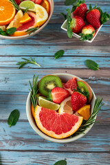 Top view of bowls with fresh fruits like strawberries, kiwi, grapefruit and citrus fruits, decorated with rosemary and mint on blue rustic table. Concept of healthy eating, summer and freshness