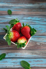 Fresh strawberries in square bowl on rustic blue wood background, decorated with mint leaves. Copy space. Concept of healthy eating, freshness and natural diet