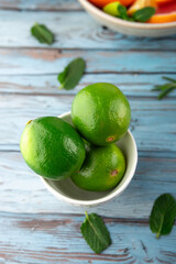 Close-up of green limes in white bowl, with citrus and mint background on blue rustic surface. Concept of natural ingredients and refreshing drinks