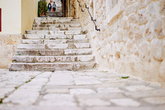 A narrow stone staircase leads up through a quaint alleyway, flanked by rustic stone walls. The steps are uneven and weathered, suggesting age. In the background, two figures are seen walking up 
