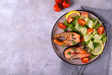 Baked trout steaks served with fresh cucumber, tomato and lettuce salad on ceramic plate on grey background. Healthy eating. Top view. Copy space