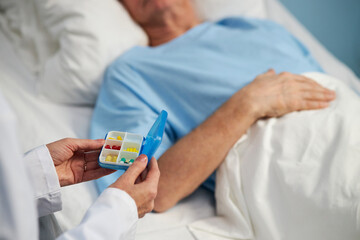 Doctor holding a pill organizer with various medications near a senior patient lying in hospital...