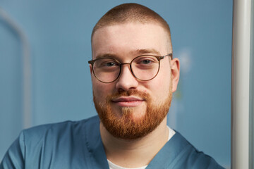 Fototapeta premium Portrait of young male medical professional smiling while looking at camera, wearing glasses and medical attire with light blue background and framed environment