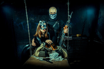 Woman, dressed in a sleek black leather outfit and wearing a cat-like mask sitting beside a duffel bag with gold bars and dollar bills. Behind her, a muscular man in black mask grips an AK-47