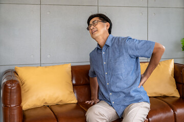 Asian elderly man experiencing lower back pain while getting up from leather sofa