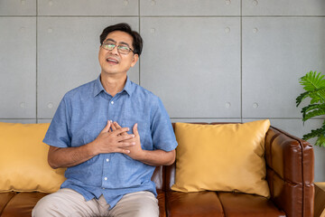 Asian elderly man experiencing chest pain, potential heart attack, sitting on a brown leather sofa