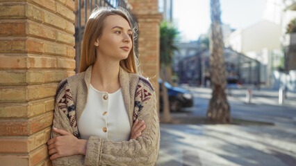 Fototapeta premium Woman standing with crossed arms on urban street, wearing cozy cardigan and looking thoughtfully into distance, sunlight highlighting her thoughtful expression and blonde hair.