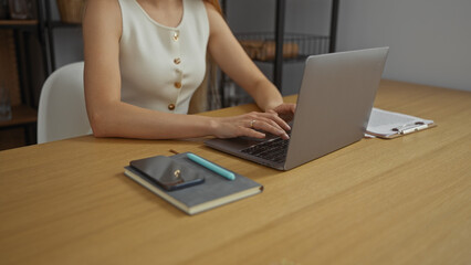 Woman typing on laptop in office setting with notebook and pen on wooden desk, conveying a professional work environment and focus on task at hand.