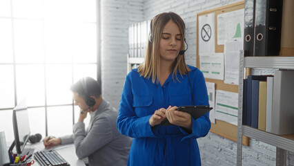 Woman in blue using tablet while man works on computer with headset in modern office, creating a focused business environment with teamwork dynamics.