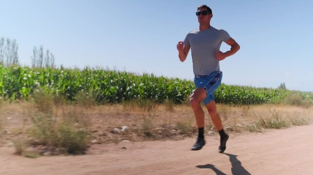 A man in athletic wear runs at a fast pace along a dusty trail by cornfields, under the scorching sun. Overcoming the heat and the challenging path, he builds endurance through intense training.