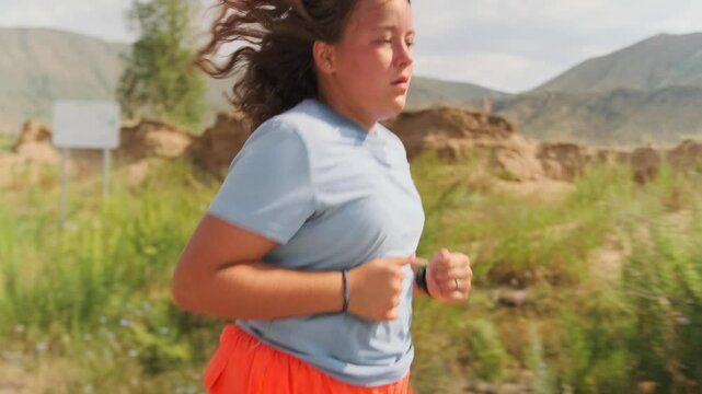 An overweight girl in athletic wear runs a scenic mountain road, the hot air making each step challenging. Pushing on through fatigue and heat, she moves closer to her goal of strength and weight loss