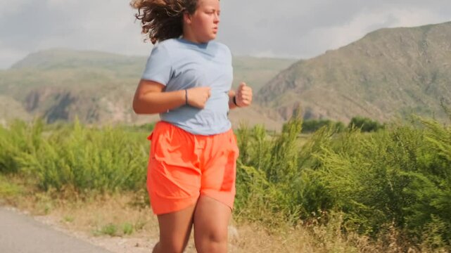 A curvy girl with loose hair in athletic wear runs along the road, battling fatigue and strain. Determined to reach her goal, she pushes forward, glancing at the storm clouds over the mountains.