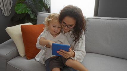 Mother and son enjoying time together indoors, sitting on a cozy sofa, sharing a tablet, surrounded by home warmth and familial love in a modern living room setting.