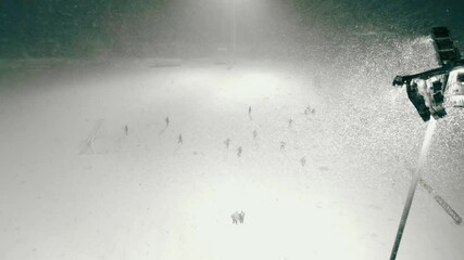Aerial drone footage showing people playing soccer or football in a snowstorm or blizzard during night or evening time while a large and bright projector light lights up the soccer field. Snowflakes. - Powered by Adobe