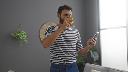 Young hispanic man indoors sipping coffee while using smartphone in a stylish living room