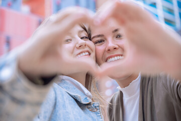 Two happy Caucasian women smiling and making a heart shape with their hands. Symbol of friendship, love, and positivity in a modern urban setting with natural light and joyful expressions.
