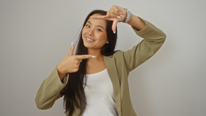 Fototapeta premium Young, attractive, chinese, woman smiling charmingly, framed by her hands, isolated against a white, background wall, bright teal blazer and white tank top, exuding confidence and elegance.