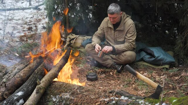 A man sits by a fire in a winter forest, sipping a hot drink near his bushcraft shelter. His backpack and gear lie nearby, creating a survival camping scene.