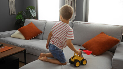 Blond toddler boy playing with a toy truck in a cozy living room with a gray sofa and orange cushions, showcasing an adorable childhood moment indoors