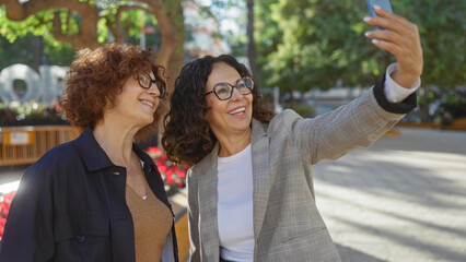 Fototapeta premium Women taking selfie in urban park setting with greenery and sunlight, showing friendship and joy in the city while enjoying outdoor time together.