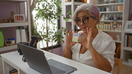 Mature woman with grey hair and glasses demonstrating a product during a videoconference against a pink background in an office setting