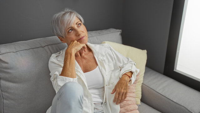 Woman relaxing on a couch in a cozy living room, with short gray hair and wearing casual clothing, looking thoughtful while being indoors in an apartment setting.