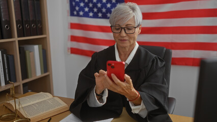Mature woman judge with short grey hair using cellphone in an office with american flag and legal...
