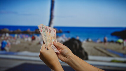 Hands holding uae dirhams at a beach setting in the background, suggesting a tropical financial or...
