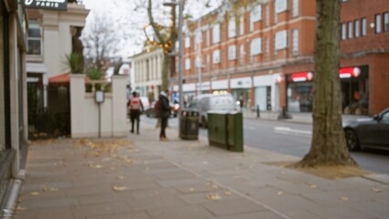 Blurred pedestrians stroll along a leaf-covered london street, capturing a defocused view of an autumn day in chelsea with urban scenery and bokeh background.