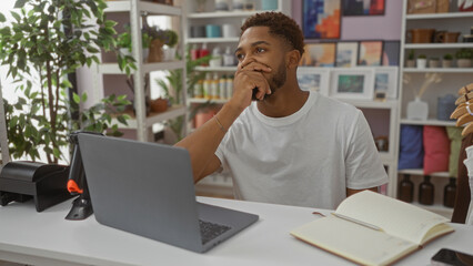 Obraz premium Young man sitting in a home decor store, thoughtfully looking at a laptop while seated at a desk with greenery and decorative items in the background