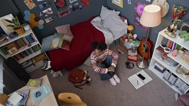 High angle full shot of sad African American gen z girl entering her room with maximalist interior, sitting on floor and feeling lonely, hugging her knees and feeling insecure