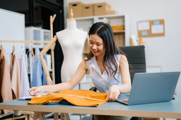  Asian young woman on desk in office of fashion designer and holds tablet, laptop and smartphone