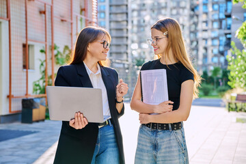 Female teacher talking to teenage student girl, outdoor, educational building background