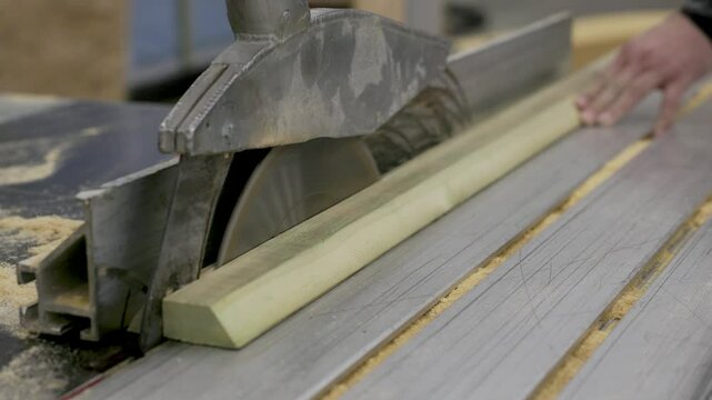 A close-up of a carpenter using a table saw to cut a wooden beam with precision. The spinning blade slices through the timber, with sawdust scattered across the surface