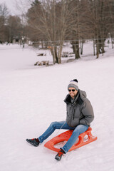 Smiling guy in sunglasses sits on a sled at the snowy edge of the forest