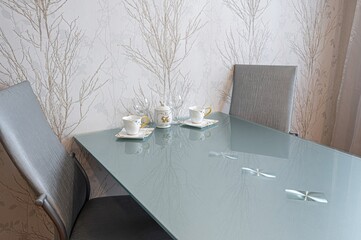 stylish dining area featuring a glass table set with teacups, wine glasses, and a sugar bowl, against patterned wallpaper.