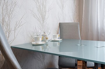 stylish dining area featuring a glass table set with teacups, wine glasses, and a sugar bowl, against patterned wallpaper.