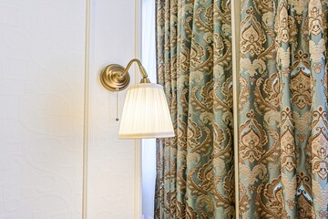 Elegant room detail: a brass wall lamp illuminating a patterned white wall, next to ornate teal and gold curtains.