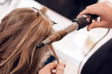 Female hairdresser straighten brown hair to pretty woman using hair iron in beauty salon.