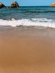 Serene coastal scene capturing golden sand, soft waves rolling ashore, and rocky formations under a clear sunny sky