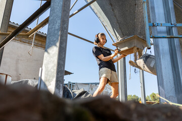 Young woman farmer is pouring organic cereals for cows from a metal silo in a farm during a sunny day, she is wearing soundproof headphones © Jordi González