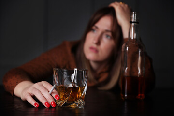 Alcohol addiction. Woman with glass of whiskey, cigarette and bottle at table indoors, selective focus