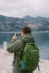 Young man with a backpack stands on the pier and takes pictures of the mountains. Back view