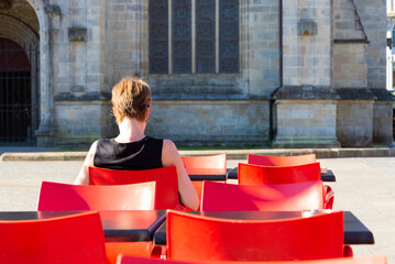 Woman sitting in a traditional cafè on a red chair, waiting for breakfast in the historical centre of Quimper, Brittany, France. Walls of the cathedral in the blurred background.